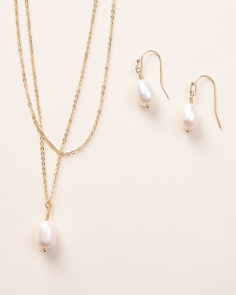 Pu&ograve; includere: Una delicata collana a catena dorata con un ciondolo a perla bianca singola, abbinata a orecchini pendenti a perla abbinati. La collana ha un design a forma di Y con due fili di catena.