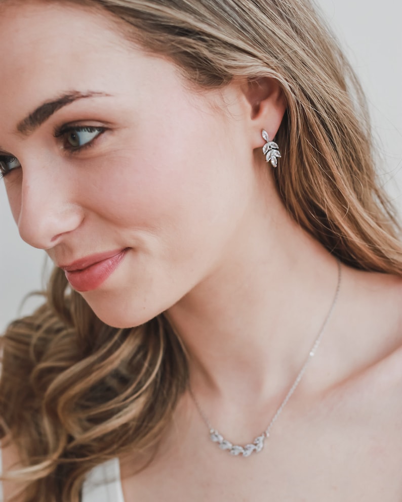 May include: A woman wearing a silver necklace and earrings with a leaf design. The necklace and earrings are made of small, clear stones.
