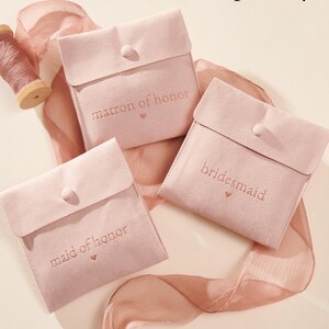 May include: Three pink velvet pouches with the words "matron of honor", "bridesmaid", and "maid of honor" embroidered on them. The pouches are arranged on a pink satin ribbon. The text "Sold separately" is at the top of the image. The text "See description for link" is at the bottom of the image.