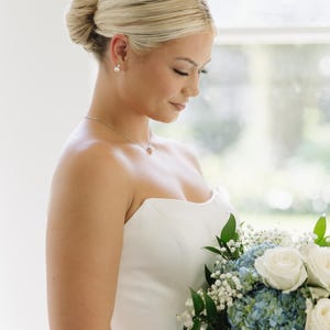 May include: A strapless white gown paired with a bouquet of white roses, blue hydrangeas, and baby's breath. The ensemble includes a delicate silver necklace with a small pendant and simple pearl stud earrings, complemented by a sleek, low bun hairstyle.
