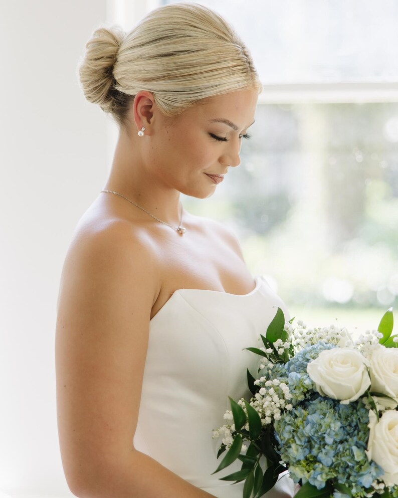 May include: A strapless white gown paired with a bouquet of white roses, blue hydrangeas, and baby's breath. The ensemble includes a delicate silver necklace with a small pendant and simple pearl stud earrings, complemented by a sleek, low bun hairstyle.