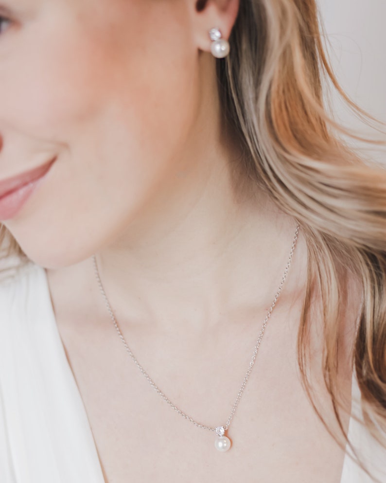 May include: A silver-toned necklace featuring a single white pearl pendant suspended from a delicate chain, accompanied by matching stud earrings. Each earring consists of a small clear stone set above a round white pearl, creating a coordinated set.