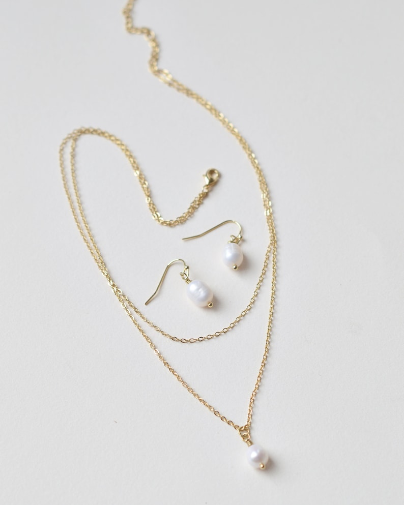 Pu&ograve; includere: Una collana a catena d'oro con due fili e un ciondolo di perla. Sono inclusi orecchini pendenti di perla abbinati.