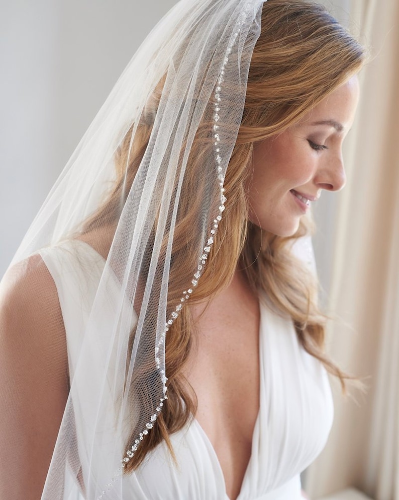 Op de afbeelding: Een witte bruids sluier met een enkele rij glinsterende versieringen langs de rand. De sluier is gemaakt van zacht tule en is perfect voor een eenvoudige en elegante bruiloft look.