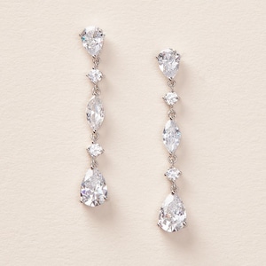 May include: A pair of elegant drop earrings. Each earring features a pear-shaped, clear gemstone at the bottom, followed by a round gemstone, a marquise-shaped gemstone, and another round gemstone, all connected by a silver chain. The earrings are set against a neutral background.