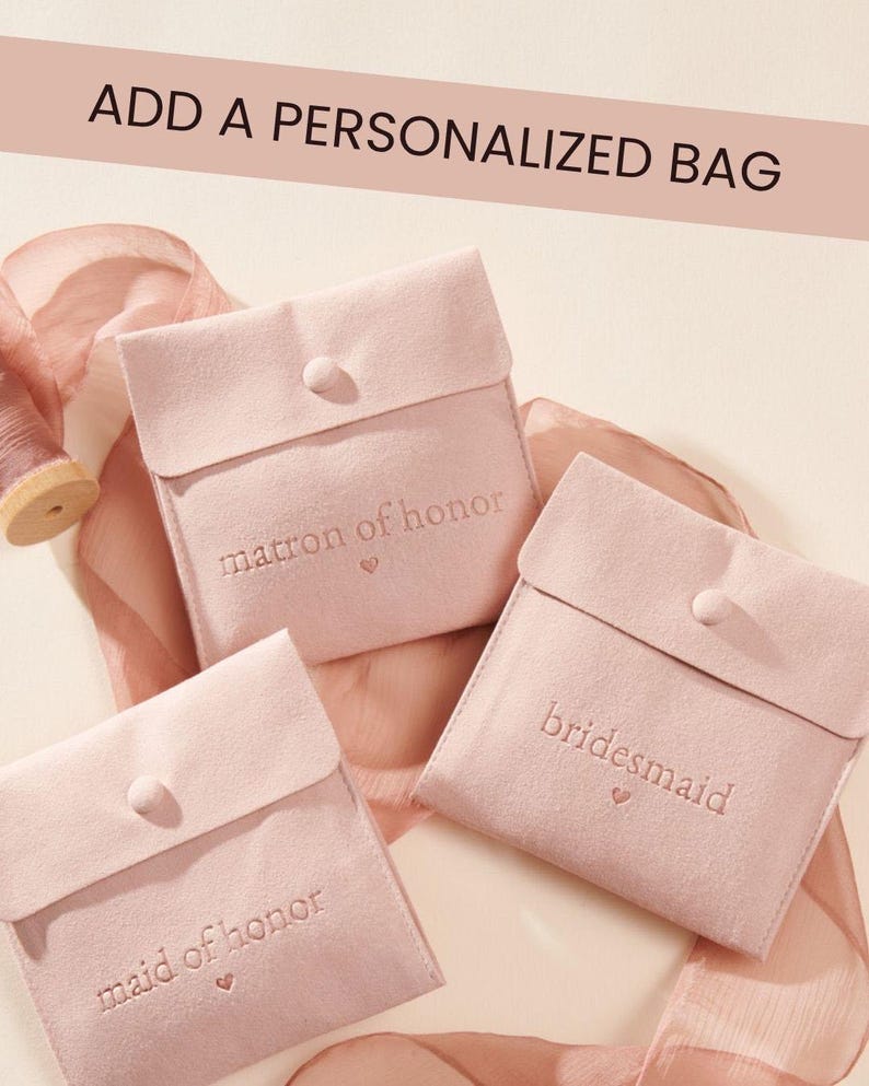 May include: Three square, light pink velvet pouches with snap closures. Each pouch features embroidered text: matron of honour, bridesmaid, and maid of honour, each with a small heart icon. The text ADD A PERSONALIZED BAG appears at the top.