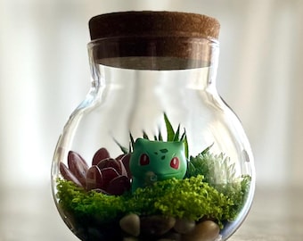 Puede incluir: Un terrario de cristal transparente con tapa de corcho que contiene una pequeña figura verde de Bulbasaur, musgo verde vibrante, hojas suculentas de color rojo oscuro y guijarros lisos en la base del recipiente redondeado.
