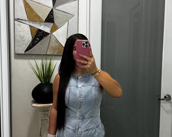 May include: A light blue denim romper featuring a sleeveless bodice, a round neckline, and a front button-down closure. The garment has a fitted waistline that transitions into a flared short bottom with visible vertical seam detailing.