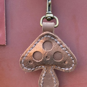 May include: A brown leather mushroom-shaped keychain featuring a metallic bronze-toned cap with circular cutouts and light-colored stitching. The charm is attached to a brass-colored swivel lobster clasp hook, hanging against a smooth brown leather surface.