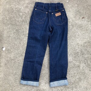 May include: A pair of dark blue denim jeans with a cuffed hem. The jeans have a brown leather patch on the back with the Wrangler logo.