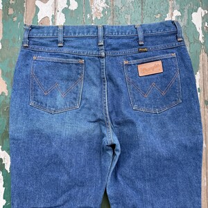 以下が含まれることがあります： バックポケットにWranglerのロゴが入った、青いデニムジーンズのクローズアップ。ジーンズは、ストレートレッグカットのクラシックな5ポケットスタイルです。ステッチは対照的なオレンジ色です。
