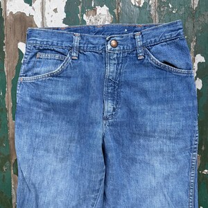 May include: Close-up of blue denim jeans. The jeans have a button closure, front pockets, and belt loops. The denim has a faded appearance, suggesting wear. The jeans are likely vintage or pre-owned.