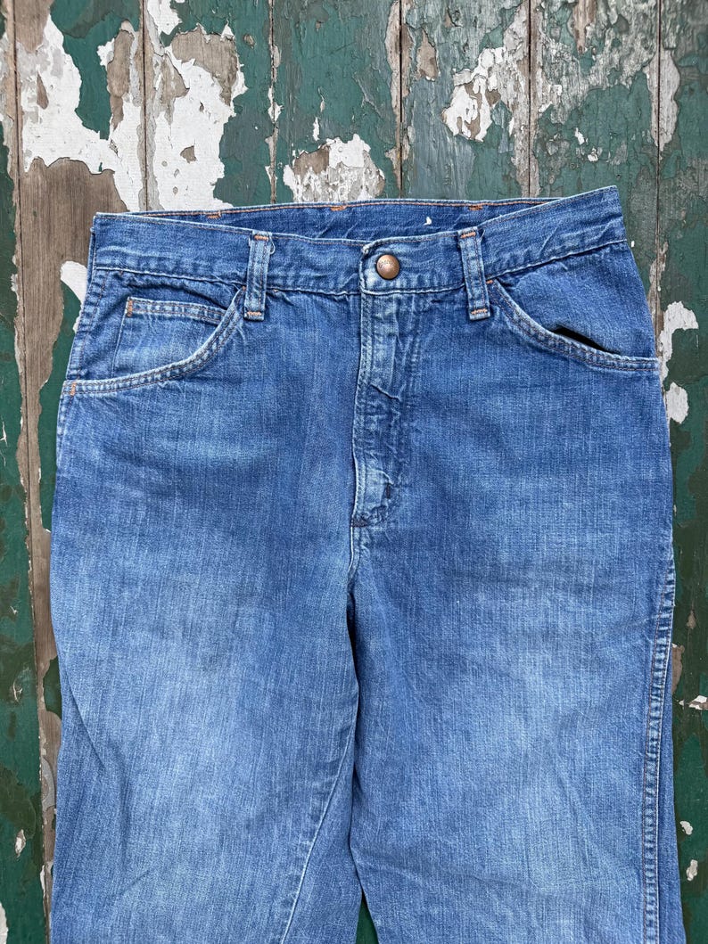 May include: Close-up of blue denim jeans. The jeans have a button closure, front pockets, and belt loops. The denim has a faded appearance, suggesting wear. The jeans are likely vintage or pre-owned.