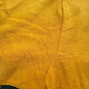 Puede incluir: Primer plano de una camiseta amarilla mostaza de manga corta. La tela muestra signos de desgaste, con textura visible y ligera decoloraci&oacute;n. La costura de la manga y el hombro es visible.