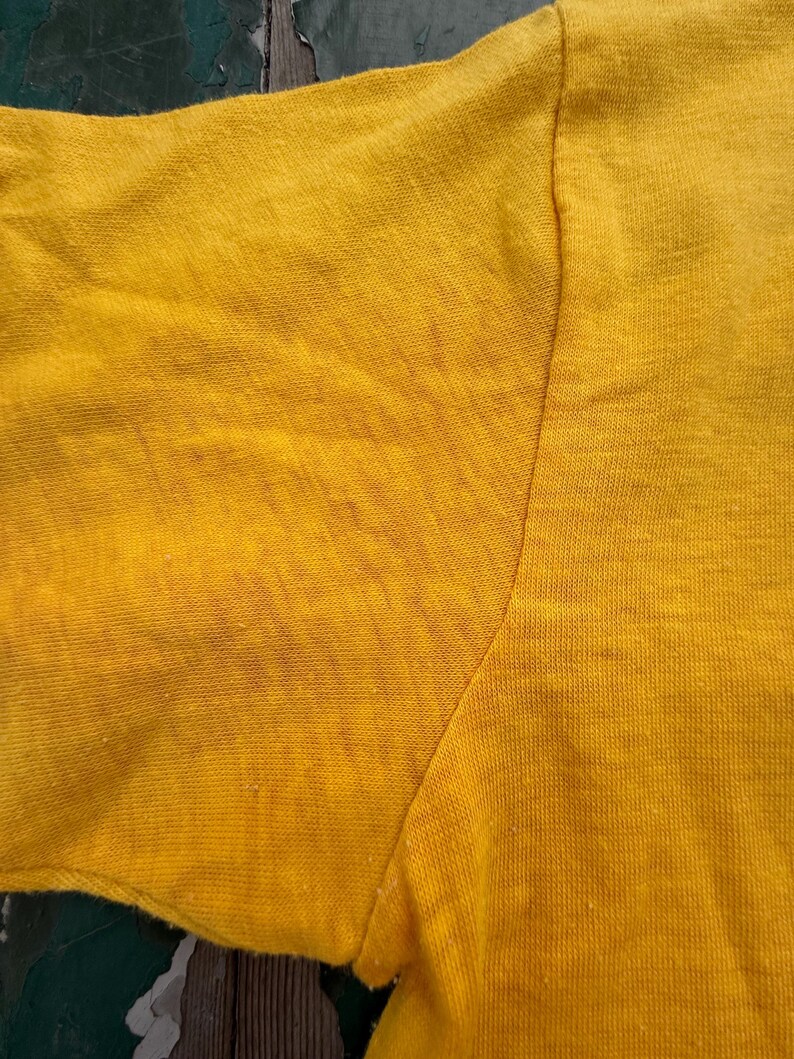 Puede incluir: Primer plano de una camiseta amarilla mostaza de manga corta. La tela muestra signos de desgaste, con textura visible y ligera decoloraci&oacute;n. La costura de la manga y el hombro es visible.