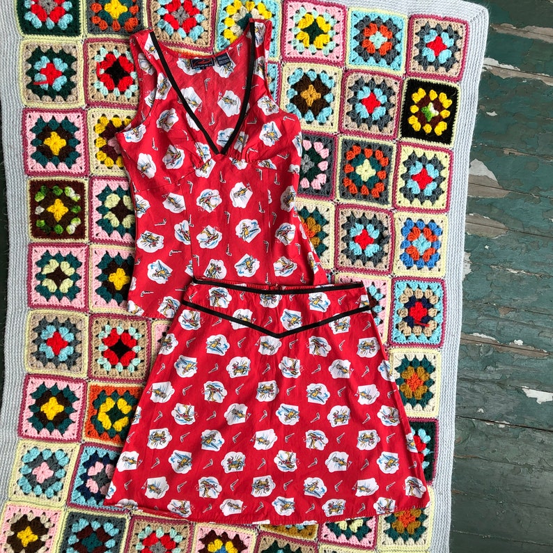 Peut inclure: Un haut sans manches rouge et blanc et une jupe assortie avec une bordure noire. Le haut a un col en V et la jupe a une taille haute. Le tissu a un motif de petits avions rouges.