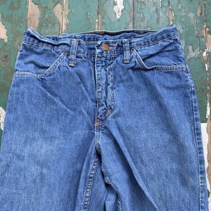 Op de afbeelding: Close-up van een blauwe spijkerbroek. De jeans heeft een knoop- en ritssluiting, voorzakken en riemlussen. De denim heeft een vervaagde uitstraling, met zichtbare stiksels in oranje draad.