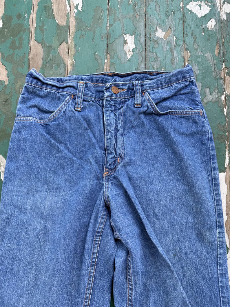 Op de afbeelding: Close-up van een blauwe spijkerbroek. De jeans heeft een knoop- en ritssluiting, voorzakken en riemlussen. De denim heeft een vervaagde uitstraling, met zichtbare stiksels in oranje draad.