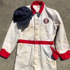 May include: White work overalls with red trim and a red and blue patch that says "Oldsmobile Service". The overalls are worn and have a few stains. A blue work cap and a red bandana are also included.