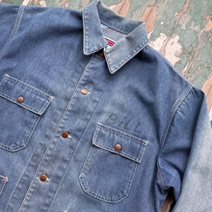 May include: A faded blue denim jacket with four front pockets and copper-colored buttons. The name "BILL" is stenciled on the right pocket. The jacket has a classic collar and is made of durable denim.