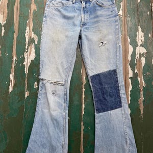 May include: Light blue denim jeans featuring a high-waisted cut, a button fly, and a classic five-pocket design. The fabric shows signs of wear with a distressed rip at the right knee and a large, dark blue rectangular patch on the left leg.