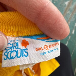 Puede incluir: Primer plano de una etiqueta de prenda amarilla con las palabras "OFFICIAL GIRL SCOUTS" en azul y blanco. El logotipo de las Girl Scouts es un tr&eacute;bol naranja. La etiqueta tambi&eacute;n dice "GIRL SCOUTS NEW YORK, N.Y."