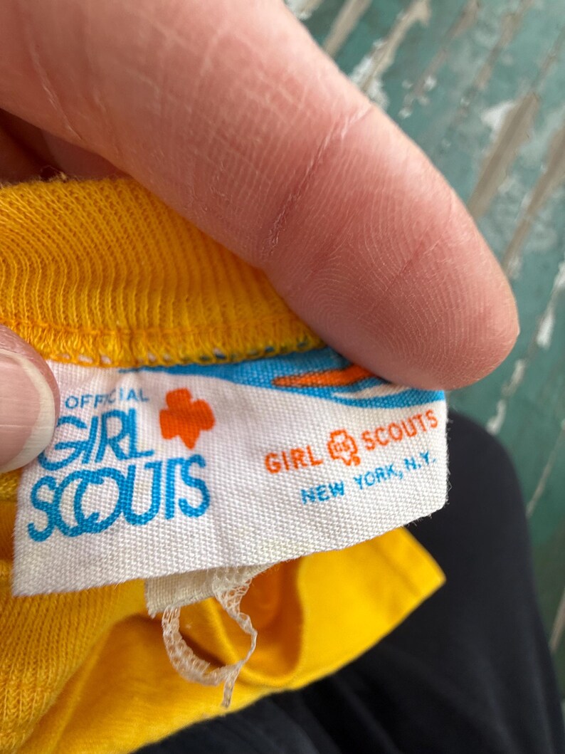 Puede incluir: Primer plano de una etiqueta de prenda amarilla con las palabras "OFFICIAL GIRL SCOUTS" en azul y blanco. El logotipo de las Girl Scouts es un tr&eacute;bol naranja. La etiqueta tambi&eacute;n dice "GIRL SCOUTS NEW YORK, N.Y."