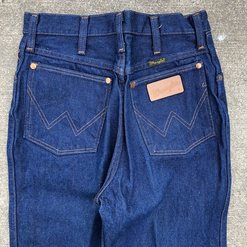 May include: A pair of dark blue denim jeans with a brown leather Wrangler label on the back pocket.