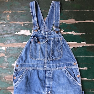 May include: Blue denim overalls with adjustable straps and multiple pockets. The overalls feature metal hardware and red stitching details. The garment is made of durable denim fabric and has a classic, vintage style.