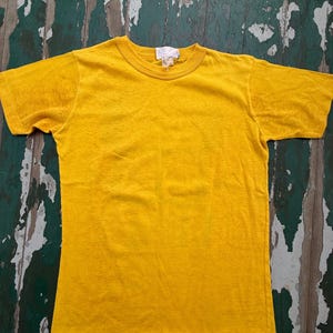 Puede incluir: Una camiseta amarilla lisa con cuello redondo. La camiseta de manga corta est&aacute; hecha de un material suave y tiene una etiqueta en el cuello. La camiseta se muestra sobre una superficie de madera desgastada.