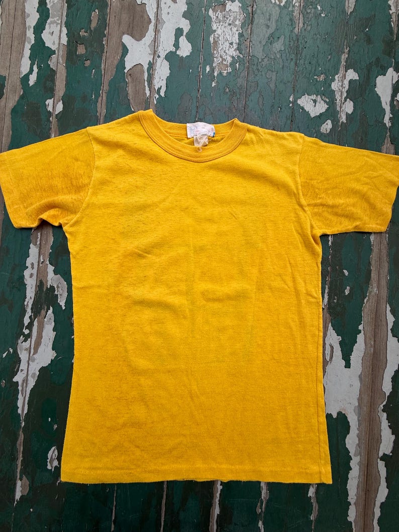 Puede incluir: Una camiseta amarilla lisa con cuello redondo. La camiseta de manga corta est&aacute; hecha de un material suave y tiene una etiqueta en el cuello. La camiseta se muestra sobre una superficie de madera desgastada.
