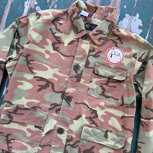 Puede incluir: Chaqueta de camuflaje en tonos marrón, verde y beige. La chaqueta tiene cierre de botones, cuatro bolsillos delanteros y un pequeño parche con un personaje de dibujos animados. La chaqueta es de estilo casual.
