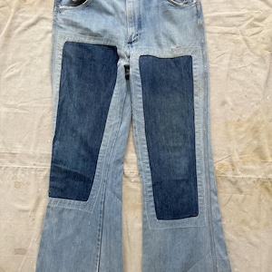 Op de afbeelding: Lichtblauwe jeans met donkerblauwe patches op de voorkant van de benen. De jeans hebben een klassiek ontwerp met vijf zakken, een knoop- en ritssluiting en een uitlopende pijpopening. De onderste zomen zijn gerafeld.