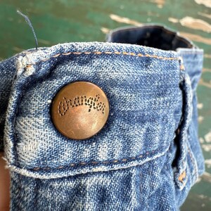 May include: Close-up of a denim garment with a copper-colored button. The button is embossed with the word "Wrangler" in a circular design. The denim is a faded blue with visible stitching.