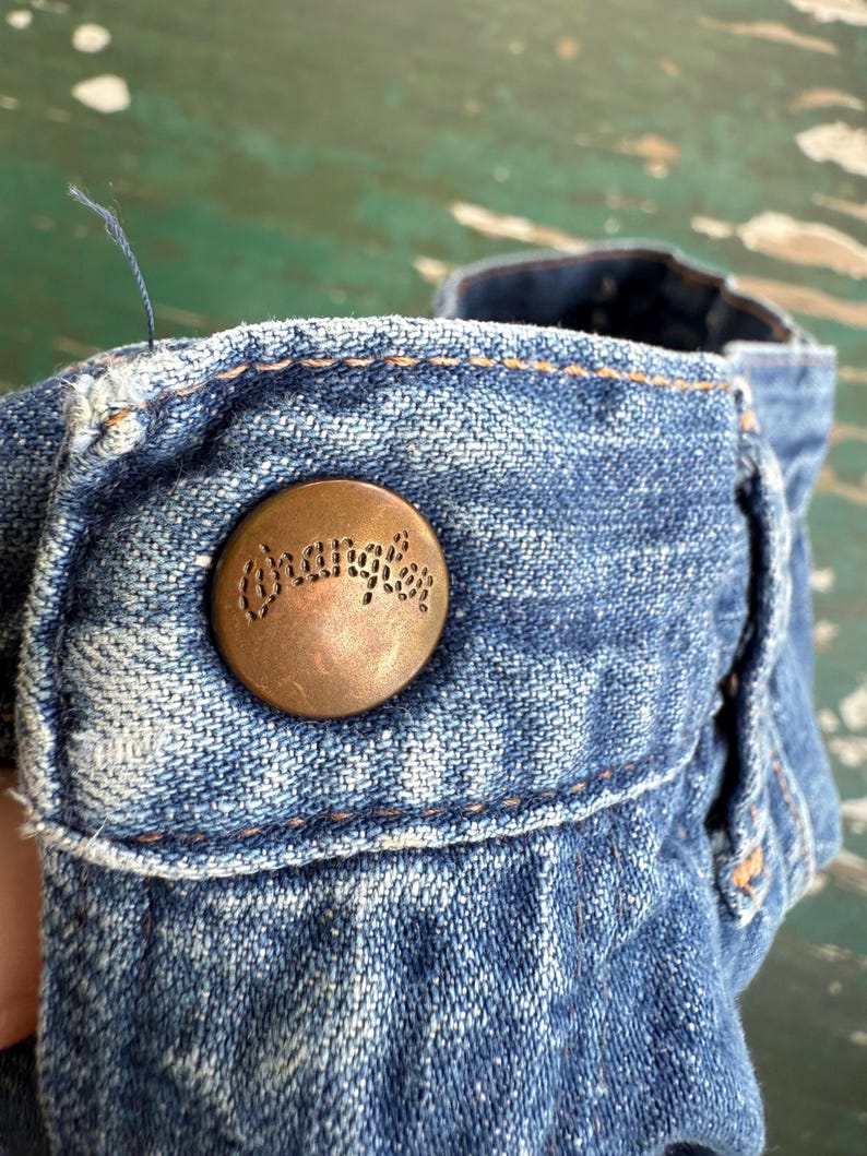 May include: Close-up of a denim garment with a copper-colored button. The button is embossed with the word "Wrangler" in a circular design. The denim is a faded blue with visible stitching.