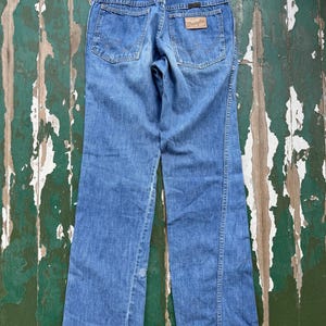 May include: Light blue denim jeans with a classic five-pocket design. The jeans feature a brown leather patch with the "Wrangler" logo on the back pocket. The jeans have a straight leg cut and are made of a durable denim material.