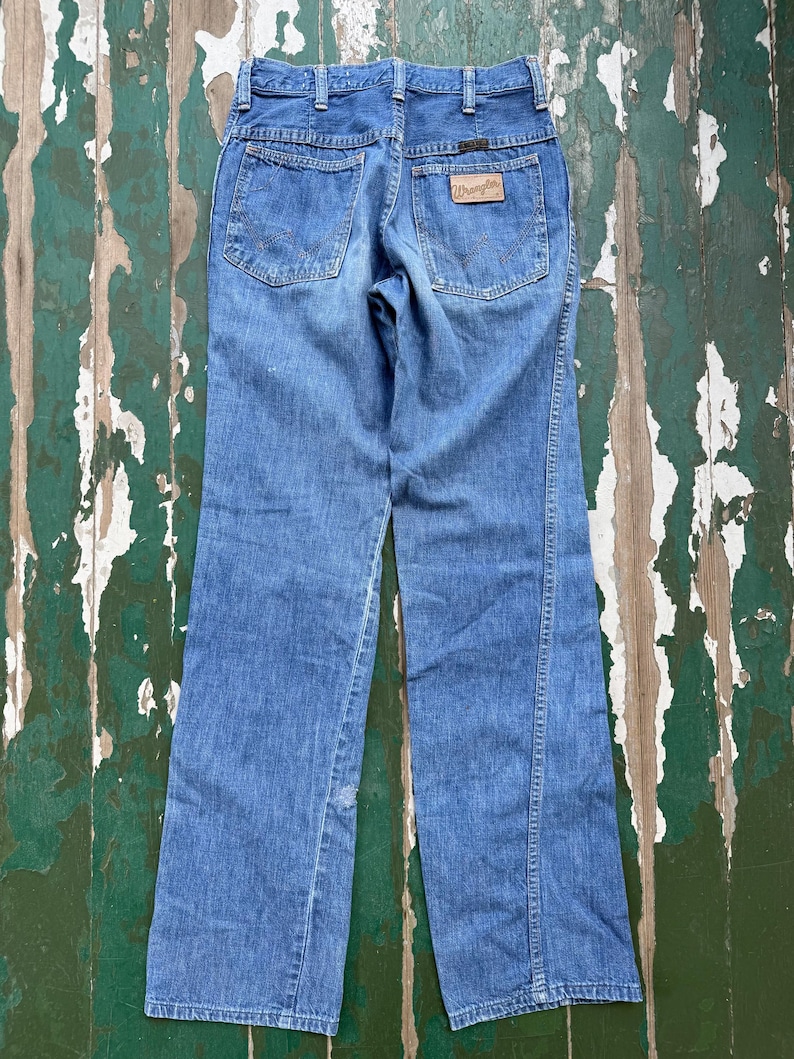 May include: Light blue denim jeans with a classic five-pocket design. The jeans feature a brown leather patch with the "Wrangler" logo on the back pocket. The jeans have a straight leg cut and are made of a durable denim material.