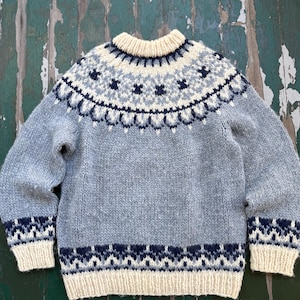 May include: A long-sleeve knit sweater featuring a light gray base with a cream-colored ribbed collar, cuffs, and hem. The yoke and lower hem area display a decorative pattern in shades of dark navy blue, light blue, and cream yarn colors.