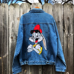 May include: A blue denim jacket with a Bugs Bunny graphic on the back. Bugs Bunny is wearing a red baseball cap, sunglasses, and a yellow shirt. He has his arms crossed.