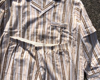 Puede incluir: Un conjunto de pijama a rayas con una camisa de manga larga y pantalones. La camisa tiene un cuello abotonado y un bolsillo en el lado izquierdo. La tela es de rayas marrones claras y blancas.