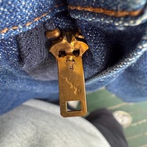以下が含まれることがあります： 青いデニム生地の真鍮製ジッパーのクローズアップ。ジッパーの引き手は長方形で、小さな刻印があります。デニムにはステッチが見え、表面は質感があります。ジッパーは部分的にかみ合っています。