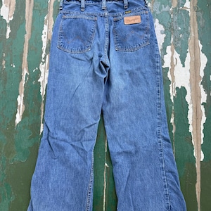 Op de afbeelding: Blauwe Wrangler jeans met een klassiek vijf-zakken ontwerp. De jeans hebben een rechte pijp en een leren patch met het Wrangler logo op de achterkant. De jeans worden getoond op een verweerd groen en wit oppervlak.