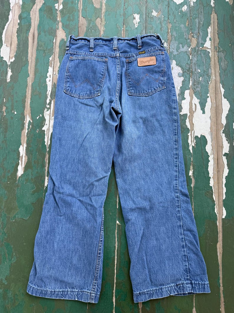 Op de afbeelding: Blauwe Wrangler jeans met een klassiek vijf-zakken ontwerp. De jeans hebben een rechte pijp en een leren patch met het Wrangler logo op de achterkant. De jeans worden getoond op een verweerd groen en wit oppervlak.