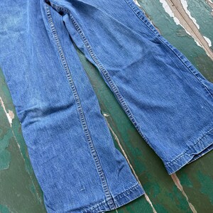 Op de afbeelding: Een spijkerbroek van blauwe denim met een klassiek ontwerp met vijf zakken. De jeans heeft een vervaagde wassing en een rechte pijp. De naden zijn versterkt met gele stiksels. De jeans wordt gepresenteerd op een verweerd houten oppervlak.