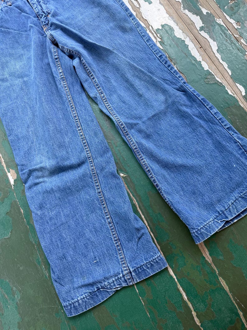Op de afbeelding: Een spijkerbroek van blauwe denim met een klassiek ontwerp met vijf zakken. De jeans heeft een vervaagde wassing en een rechte pijp. De naden zijn versterkt met gele stiksels. De jeans wordt gepresenteerd op een verweerd houten oppervlak.