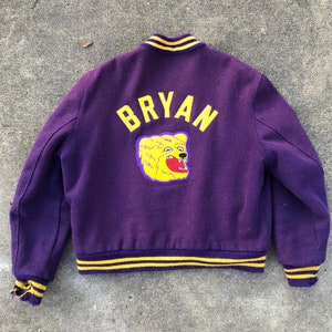 May include: Purple varsity jacket with yellow and black stripes. The jacket has the name "BRYAN" and a yellow bear head with a red mouth on the back.