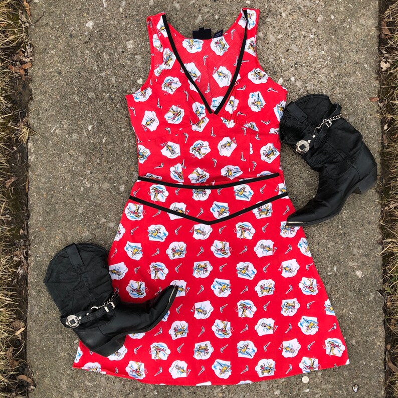 Peut inclure: Une robe rouge sans manches avec une bordure noire et un motif blanc et rouge de cowboys &agrave; cheval. La robe est associ&eacute;e &agrave; deux bottes de cowboy noires.