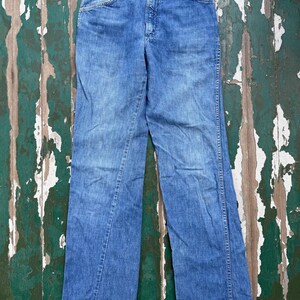 May include: A pair of light blue denim jeans with a classic five-pocket design. The jeans have a straight leg cut and a button and zipper closure. The denim has a faded appearance, suggesting wear and washing. The jeans are laid flat on a distressed wooden surface.