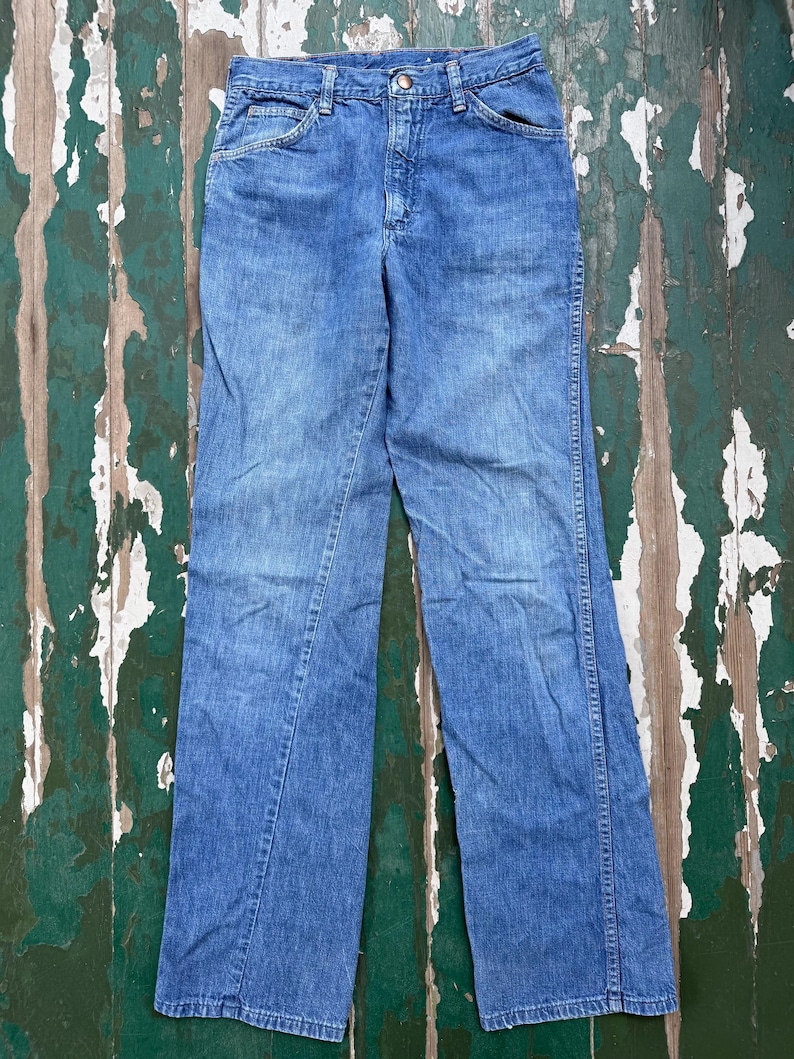 May include: A pair of light blue denim jeans with a classic five-pocket design. The jeans have a straight leg cut and a button and zipper closure. The denim has a faded appearance, suggesting wear and washing. The jeans are laid flat on a distressed wooden surface.