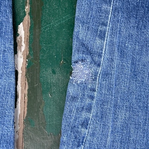 May include: Close-up of blue denim jeans with a distressed patch. The jeans show signs of wear and tear, with a visible hole and frayed edges. The background is a weathered, green-painted wooden surface.